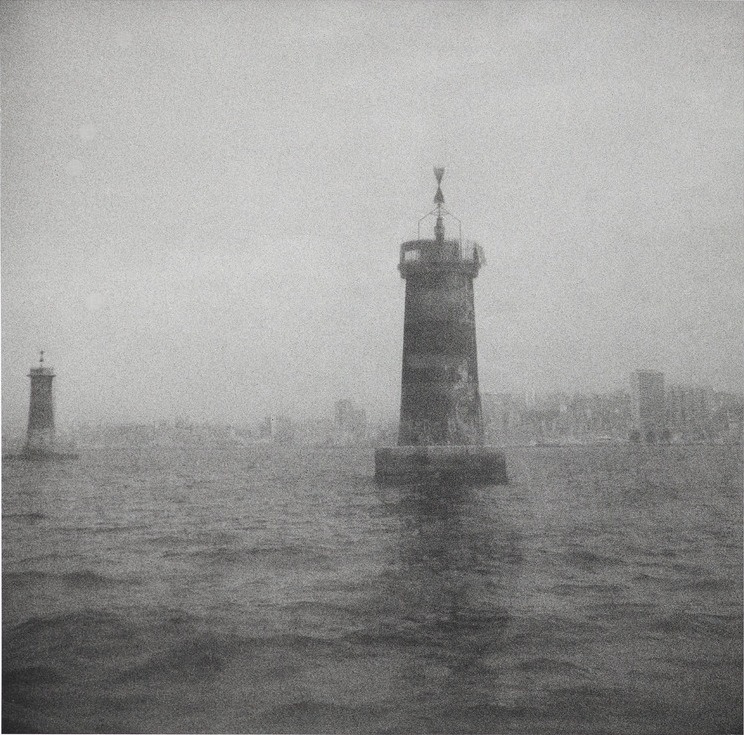 Image carrée : un phare dans la brume au milieu du port, la ville effacée derrière