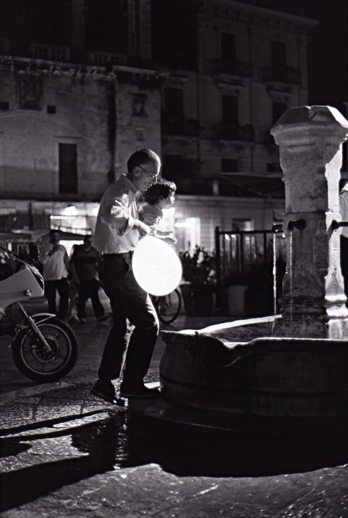 De nuit, un globe lumineux au-dessus d'une fontaine, des passants et une moto devant des façades
