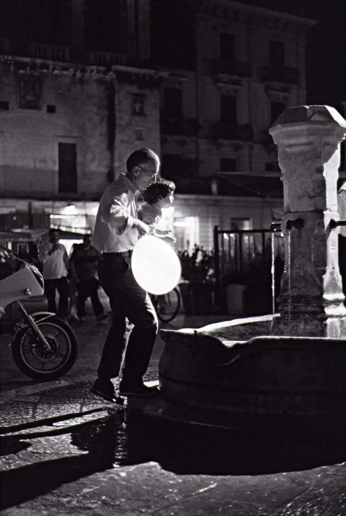 De nuit, un globe lumineux au-dessus d'une fontaine, des passants et une moto devant des façades
