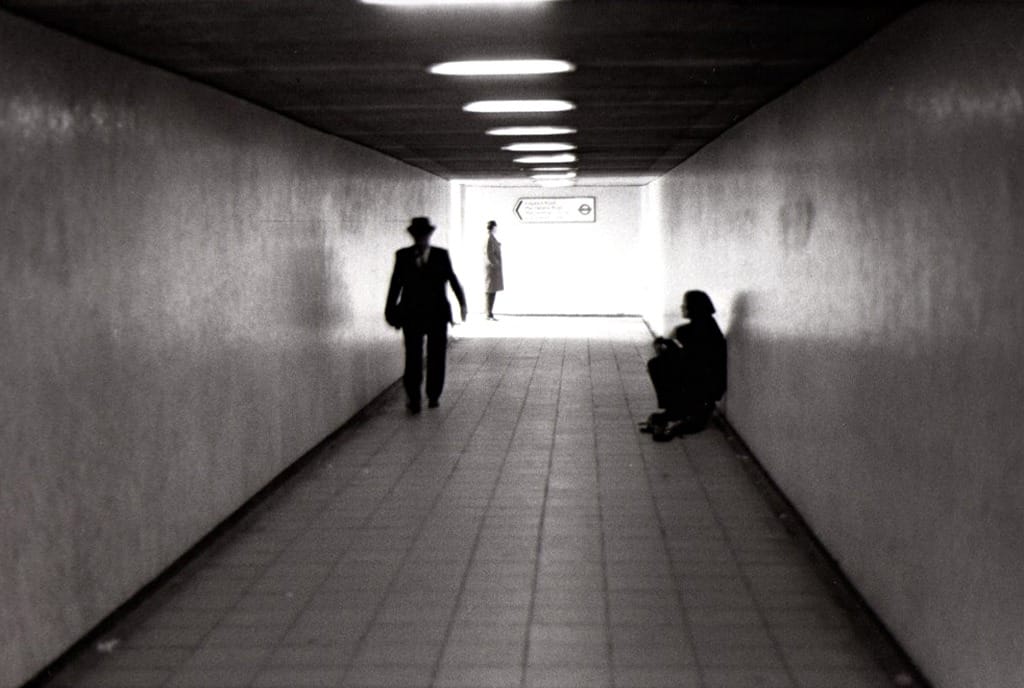 Un passage souterrain éclairé au néon, un homme qui marche vers la sortie, un autre assis contre le mur