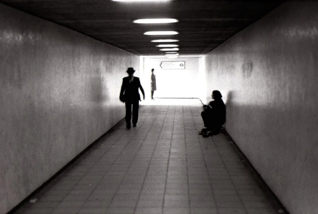 Un passage souterrain éclairé au néon, un homme qui marche vers la sortie, un autre assis contre le mur