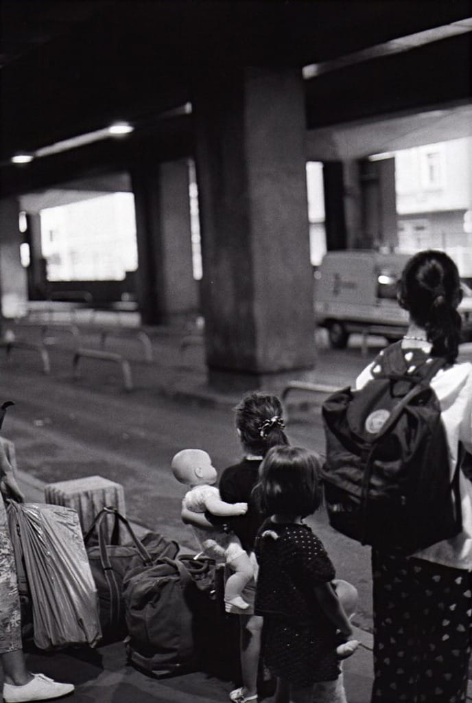 Sous un ouvrage de béton, un groupe de personnes avec sacs et enfants, en attente