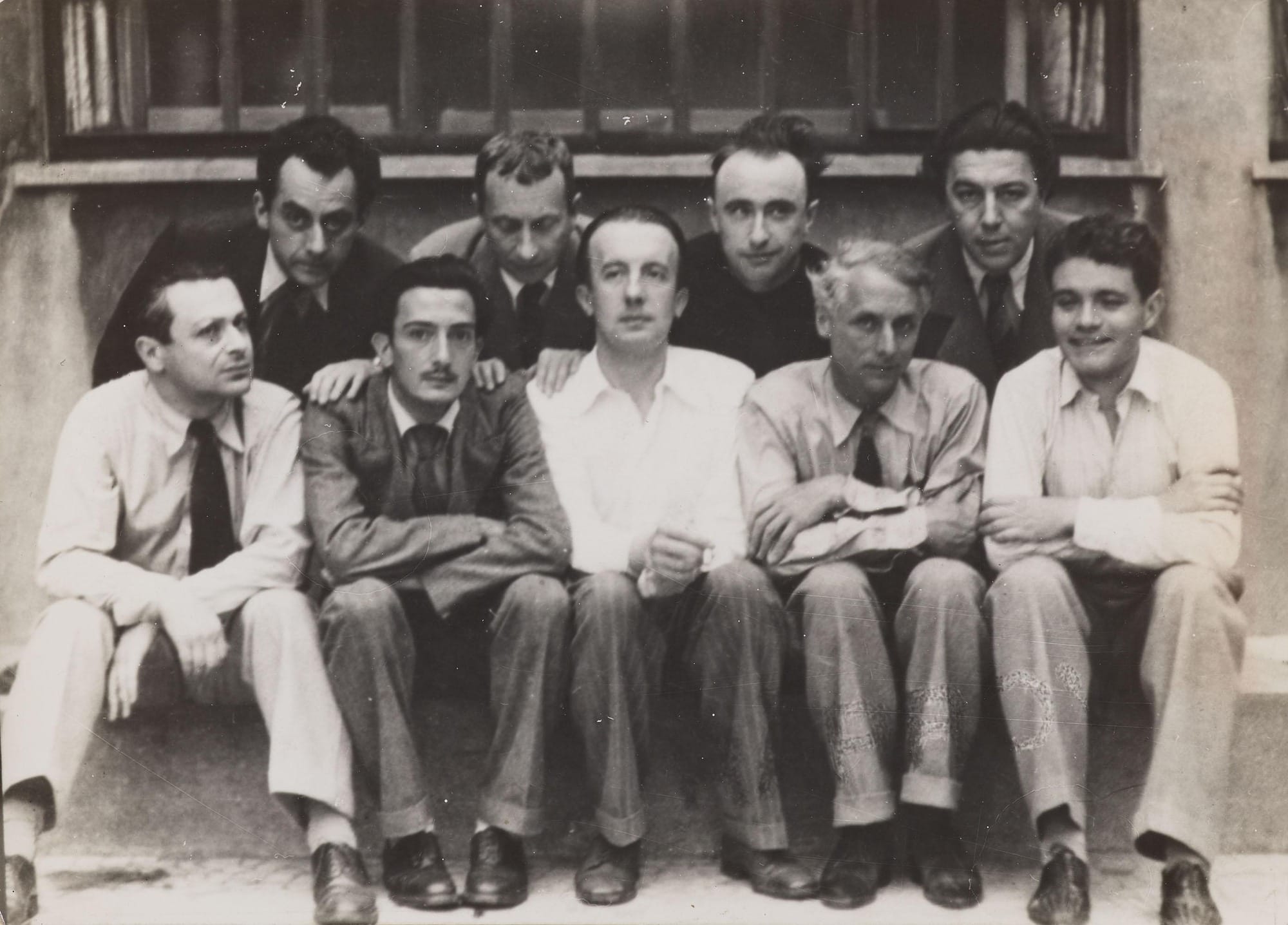 Photographie de groupe du cercle surréaliste vers 1930 : neuf hommes assis et debout devant une fenêtre