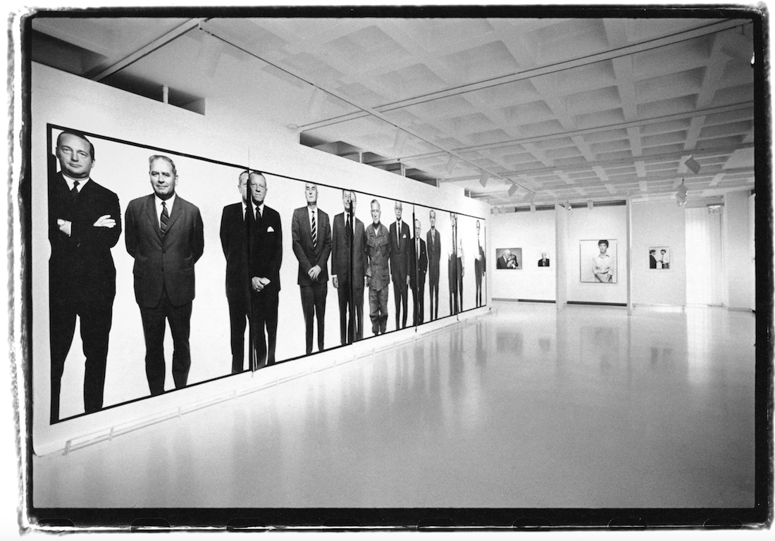 Murale de onze hommes en pied, en costume, sur fond blanc, dans une salle de musée